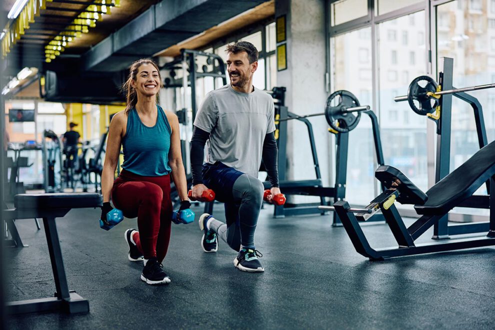 Happy athletic couple exercising with hand weights in lunge position in a gym.